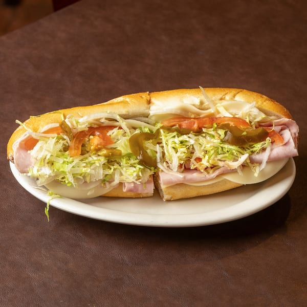 Submarine sandwich on a white plate, filled with ham, cheese, shredded lettuce, tomatoes, onions, and pickled peppers.