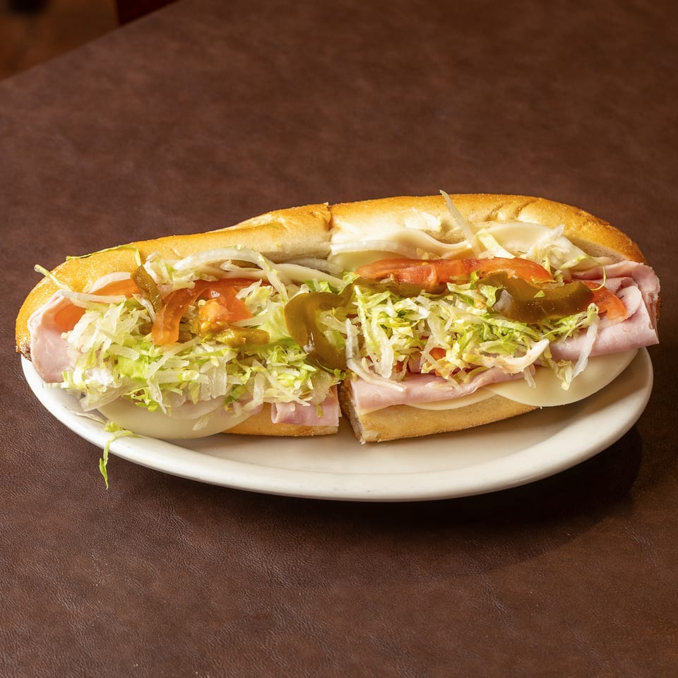 Submarine sandwich on a white plate, filled with ham, cheese, shredded lettuce, tomatoes, onions, and pickled peppers.