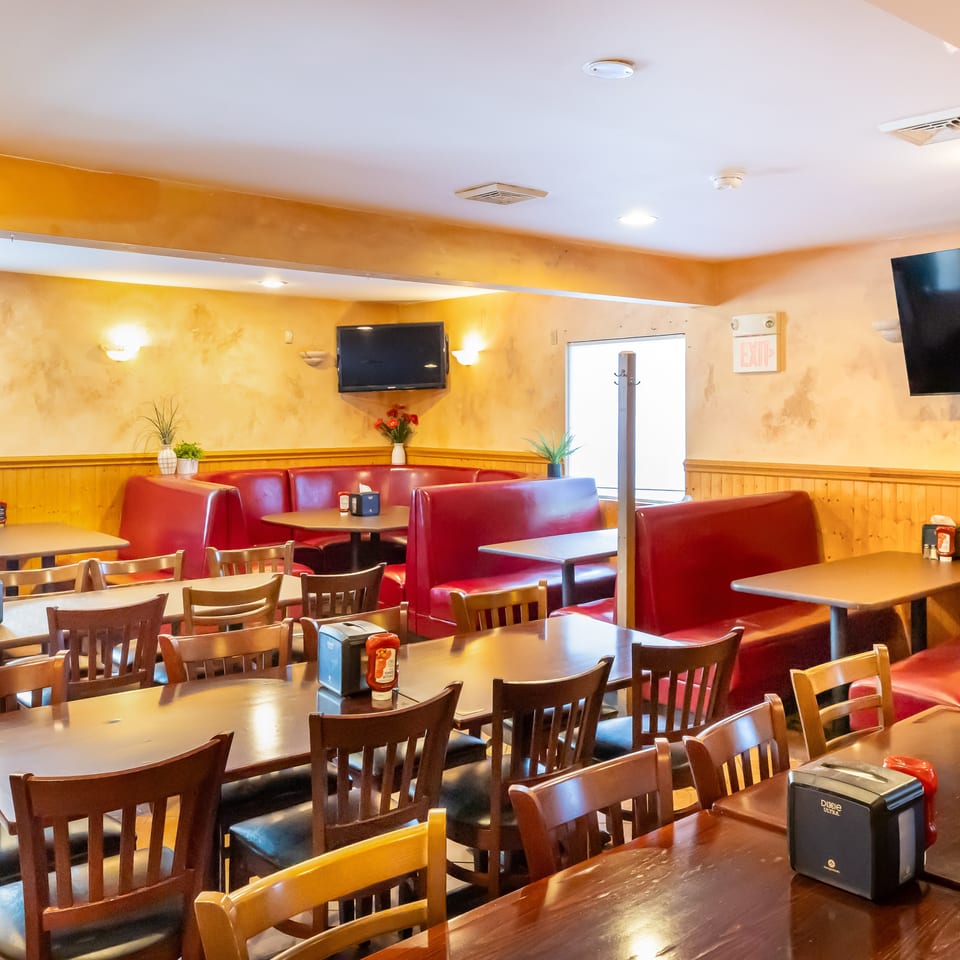 Warmly lit restaurant dining room with red leather booths, wooden tables and chairs, wall-mounted televisions, and decorative plants.