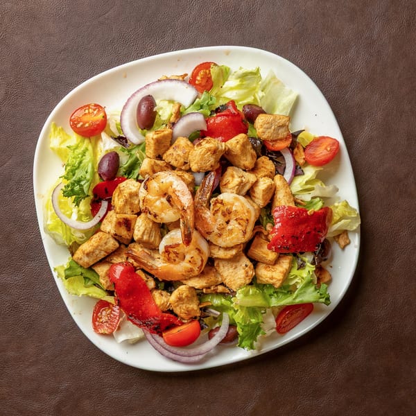Grilled chicken and shrimp salad with lettuce, tomatoes, red onion, roasted peppers, and olives on a white plate.