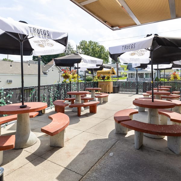 Sunny outdoor Troegs beer garden with round picnic tables, red benches, umbrellas, string lights, and hanging flower baskets.