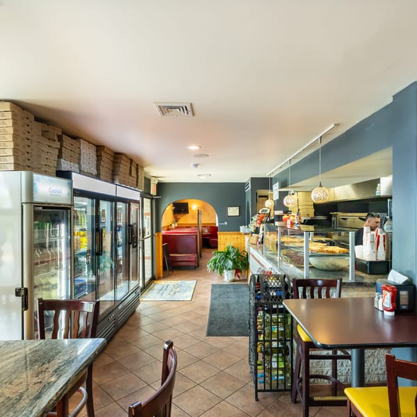 Bright casual restaurant interior with refrigerated drink cases, stacked pizza boxes, display counter, dining tables, and red booth seating.