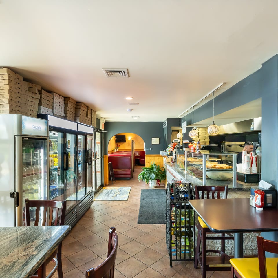 Bright casual restaurant interior with refrigerated drink cases, stacked pizza boxes, display counter, dining tables, and red booth seating.