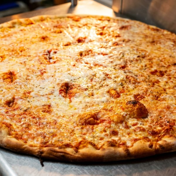 A large, freshly baked cheese pizza with a golden crust sits on a stainless steel counter.