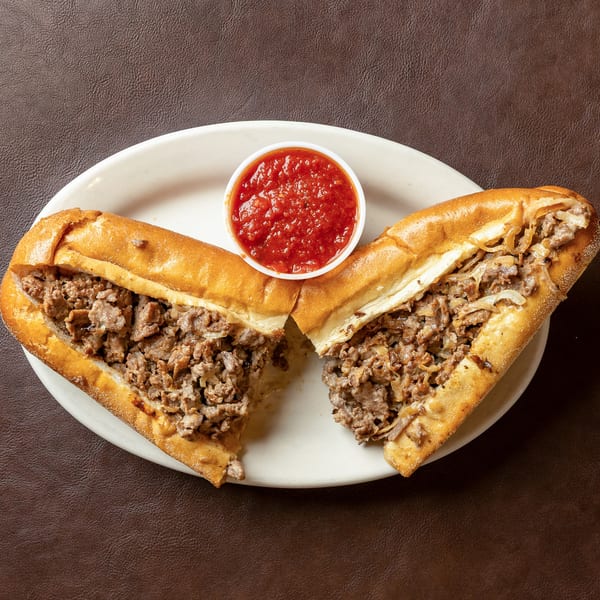 Two cheesesteak sandwiches with chopped beef and onions on a plate, served with red dipping sauce.